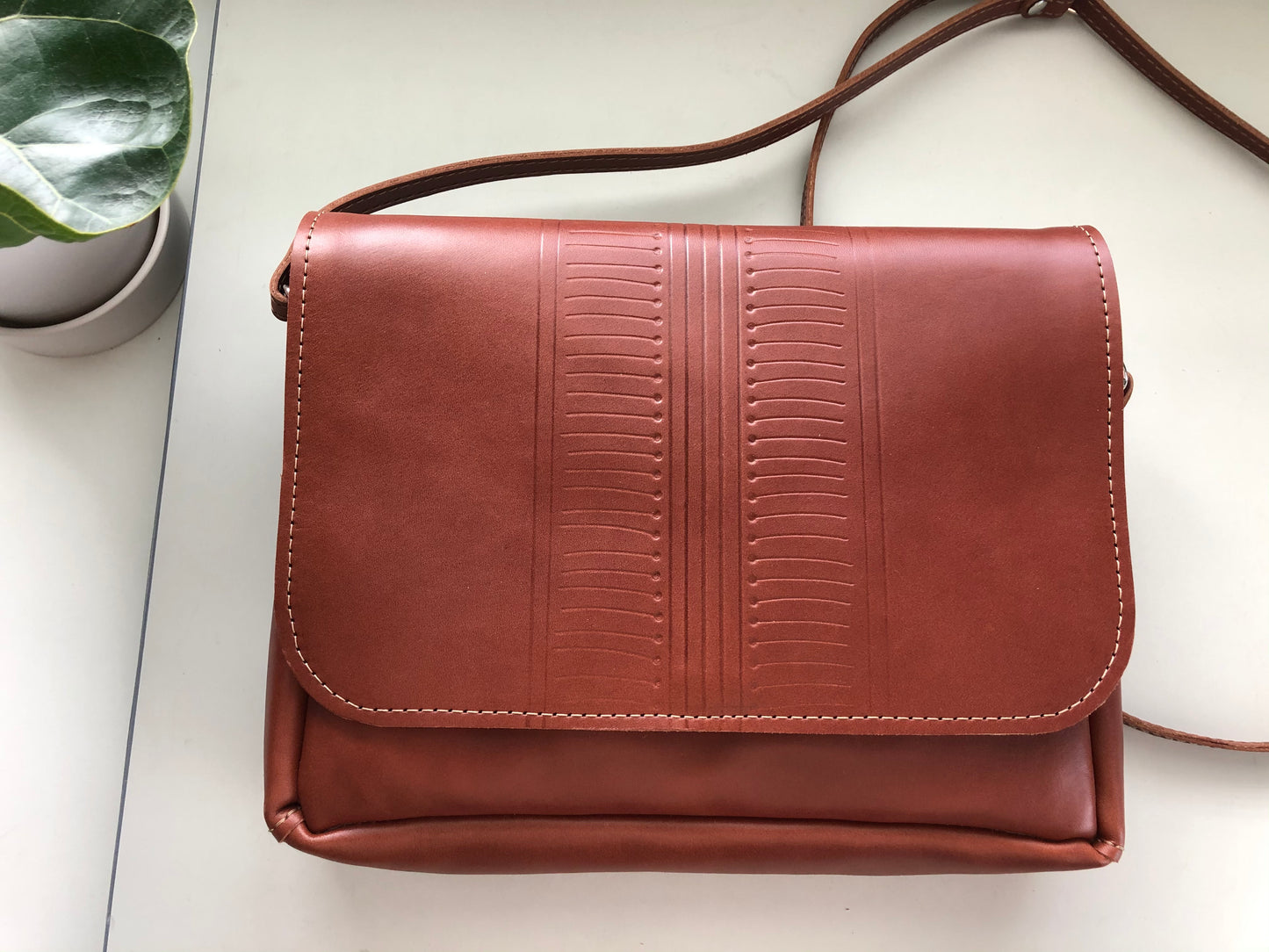 Brown leather crossbody bag lies on white table near plant.
