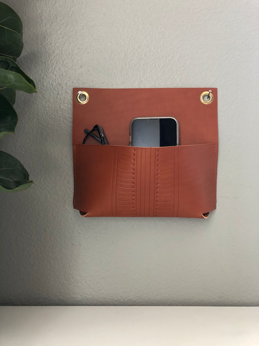 Sleek leather wall pocket holds phone and glasses near plant.