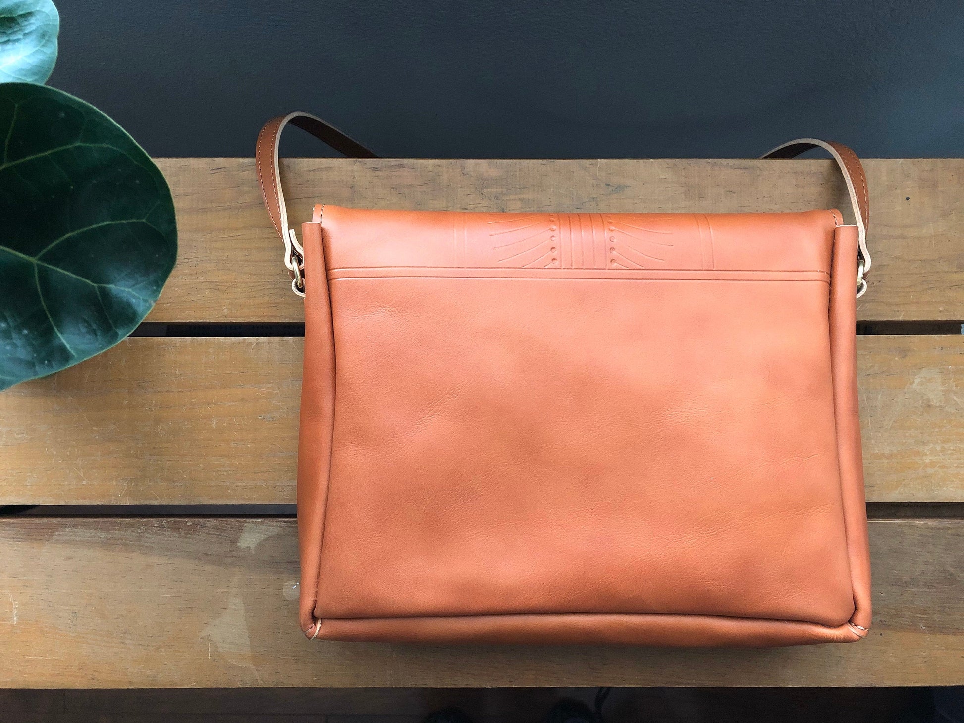 Back of tan leather crossbody bag with small tooling detail. Resting on table near plant