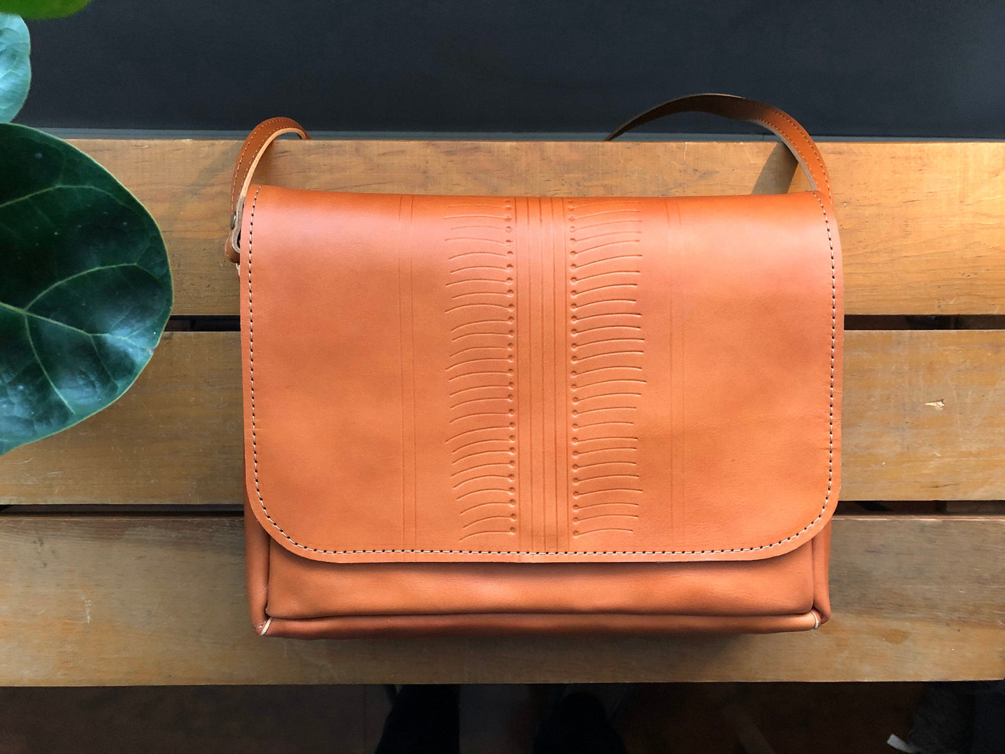 Tan leather crossbody bag with tooled design on front rests on table near plant