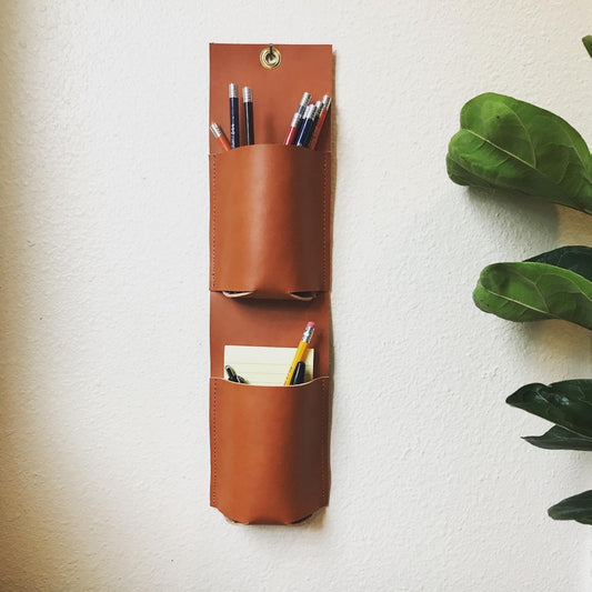 Vertical leather wall organizer with two pockets holding colored pencils, pens, and post it notes