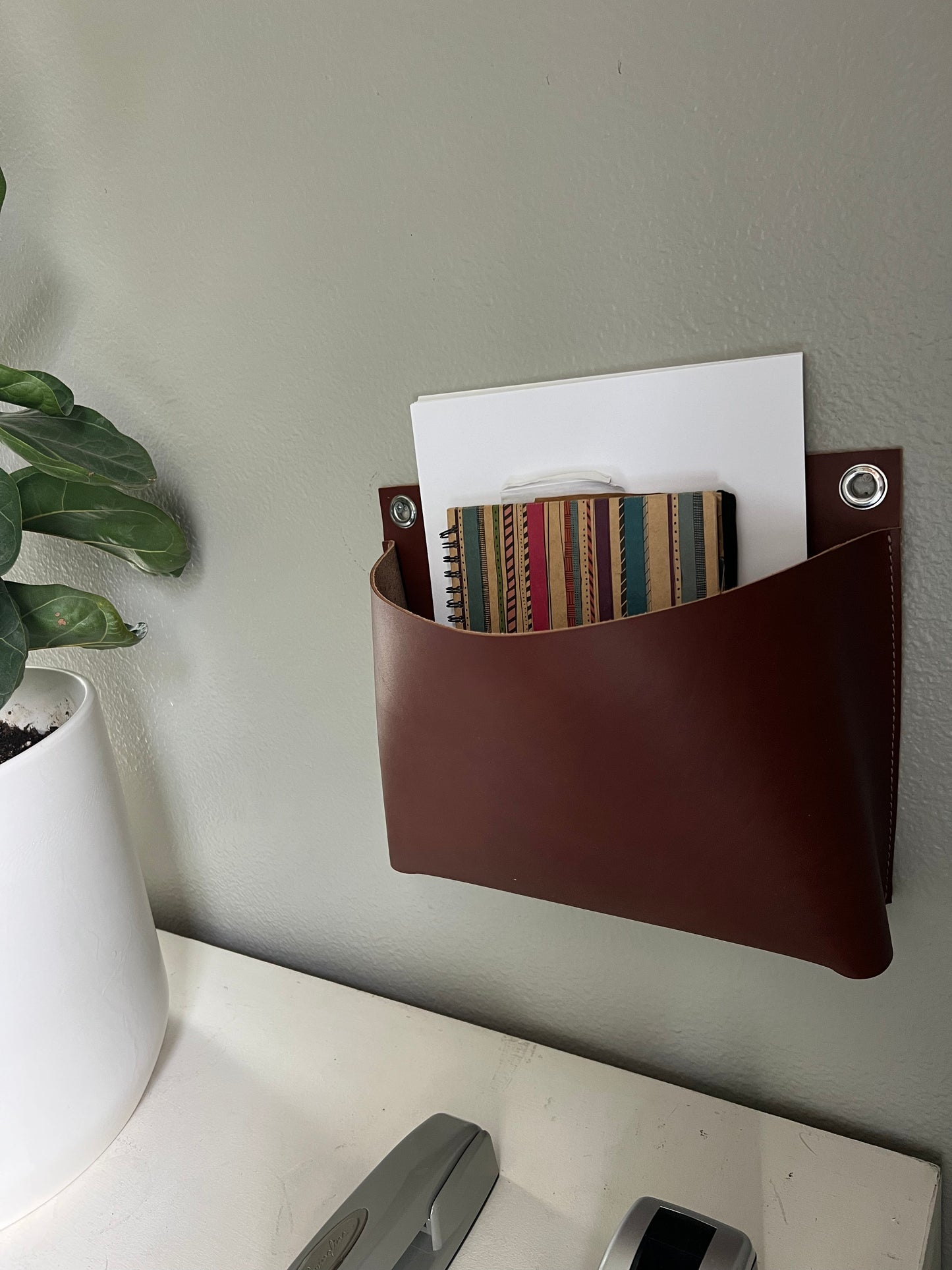 Sleek and roomy brown leather wall pocket holds a collection of papers and books near a plant.