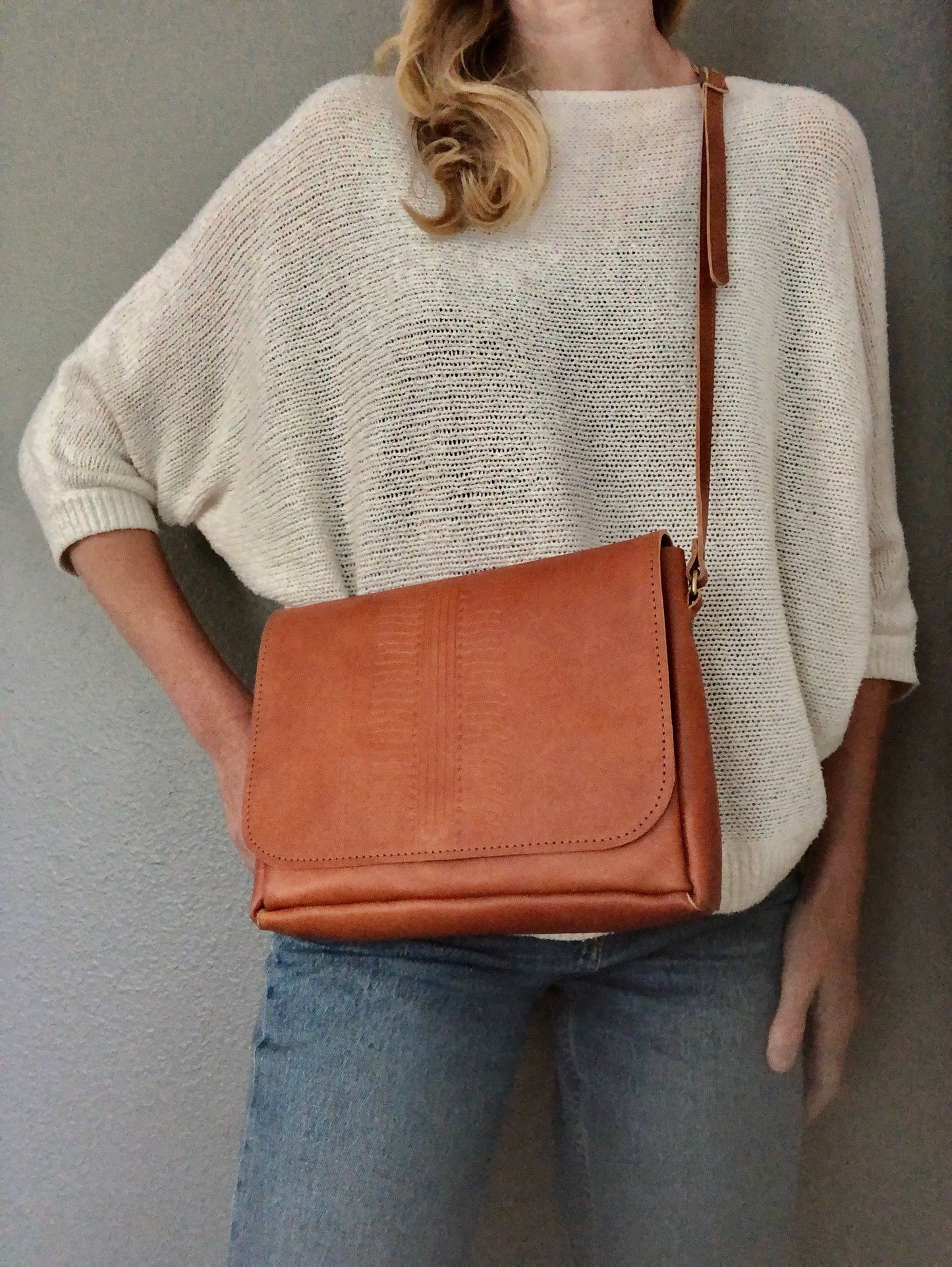 Woman poses to show buttery, tan leather purse. Purse has a subtle tooled design on the front flap.