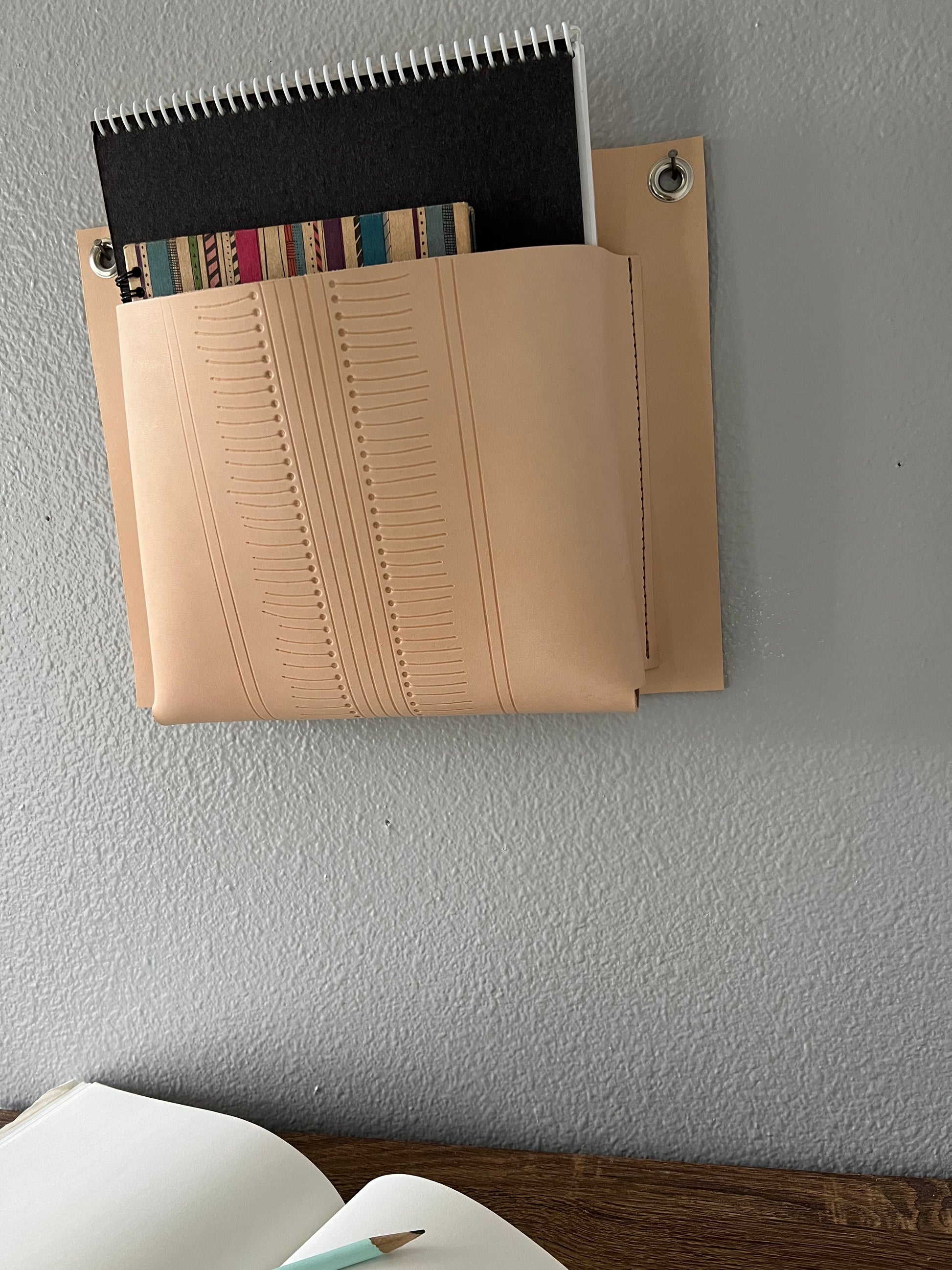 Nude leather wall pocket with geometric design, holds an assortment of sketchbooks above a desk.