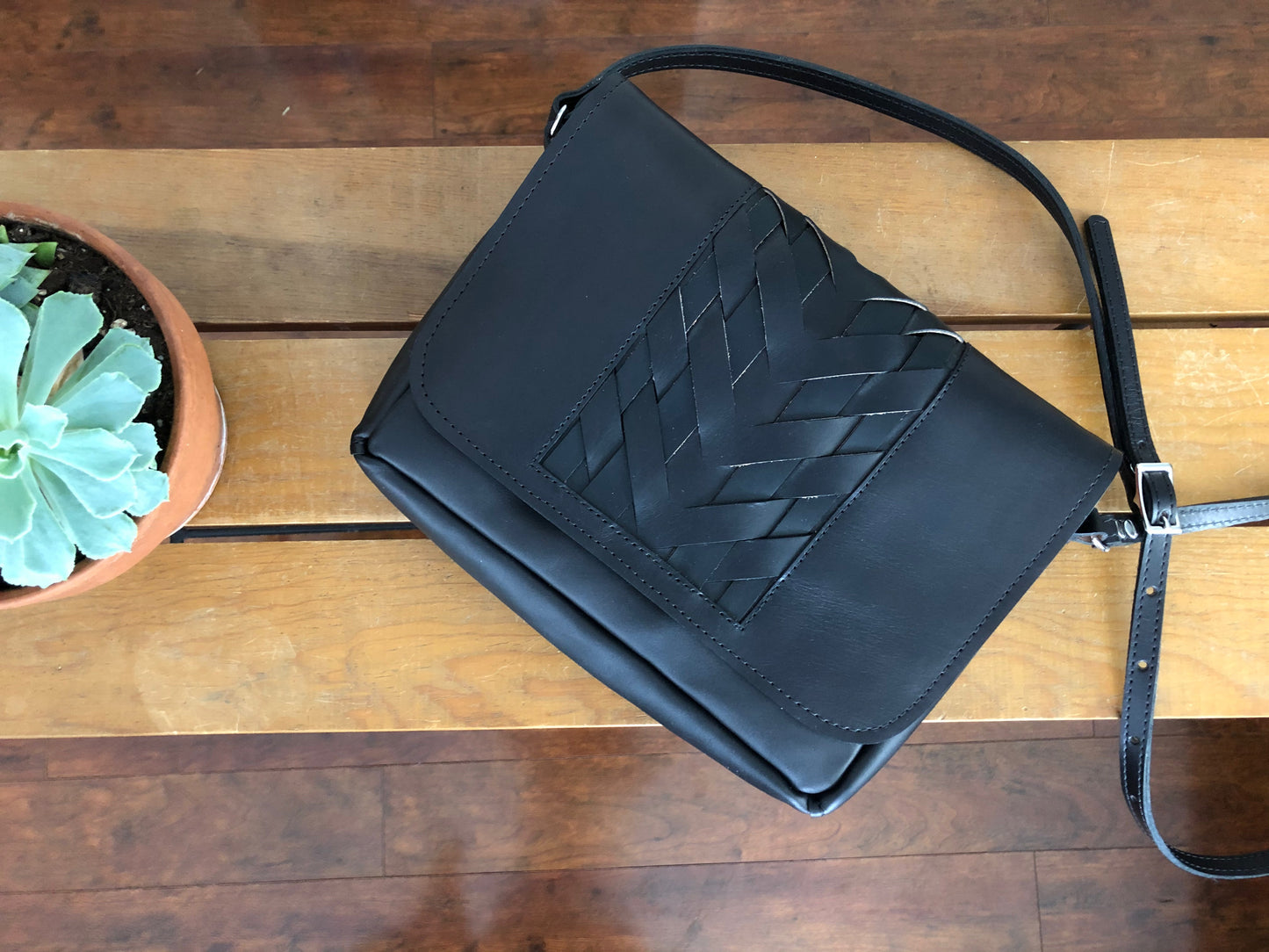 Black leather crossbody bag with woven center panel, lies on wooden table.