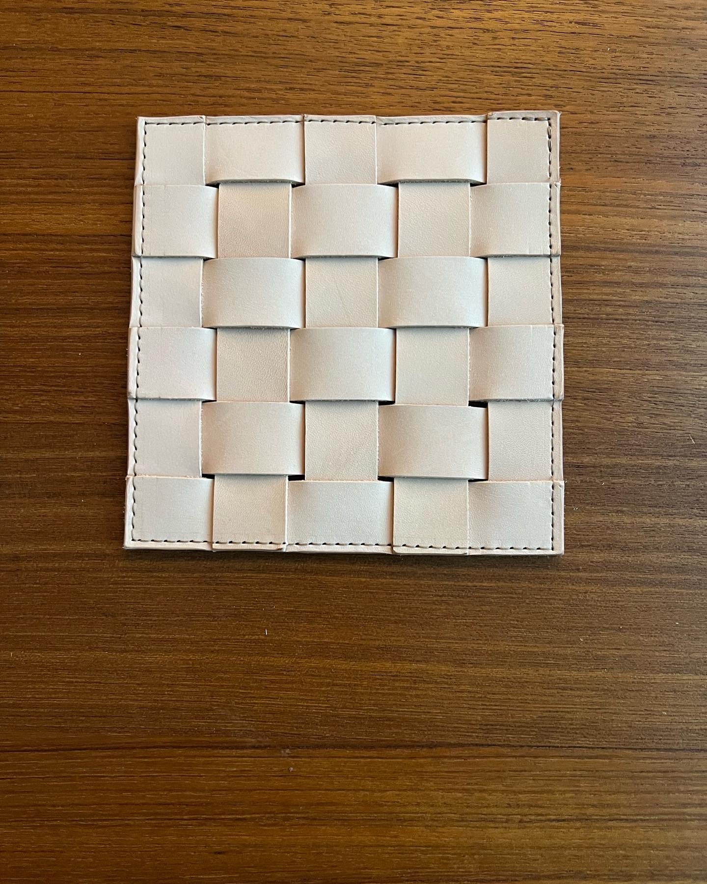 Pristine, nude leather trivet made of woven strips sits on wood table top.