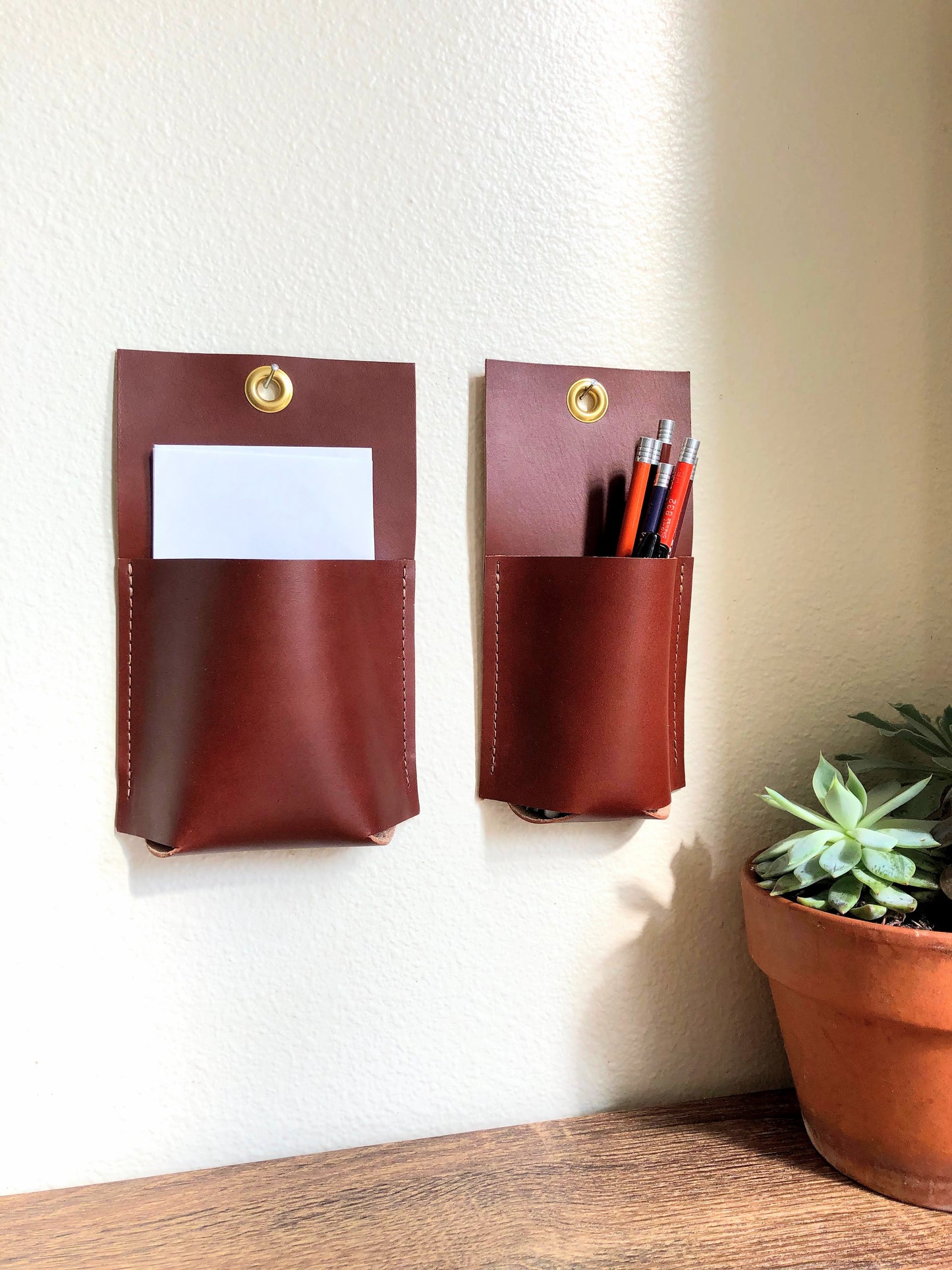 Two sizes of brown leather wall pockets are hung together near a potted plant and table.