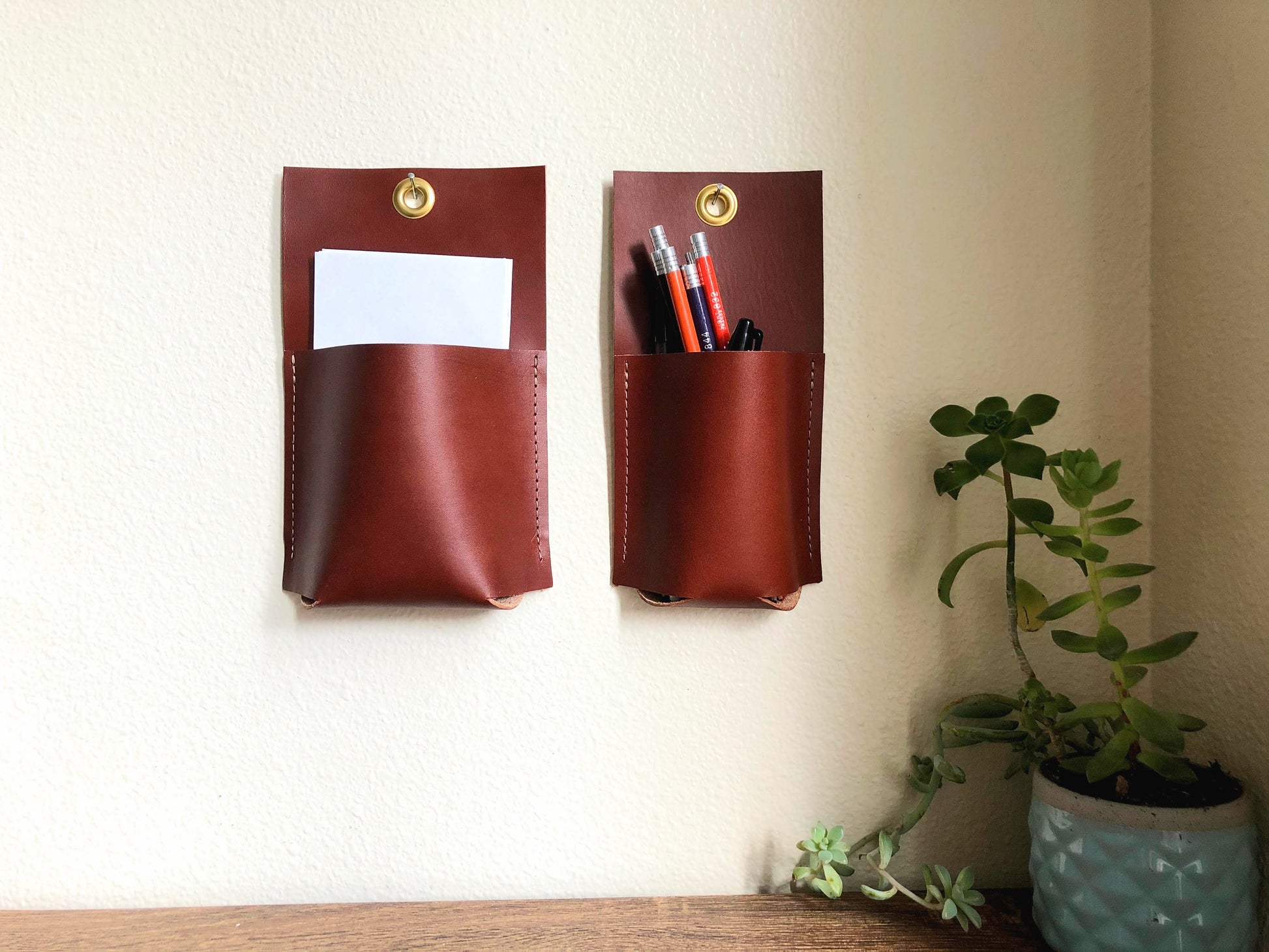 Two brown leather wall pockets hang together holding pencils and paper.