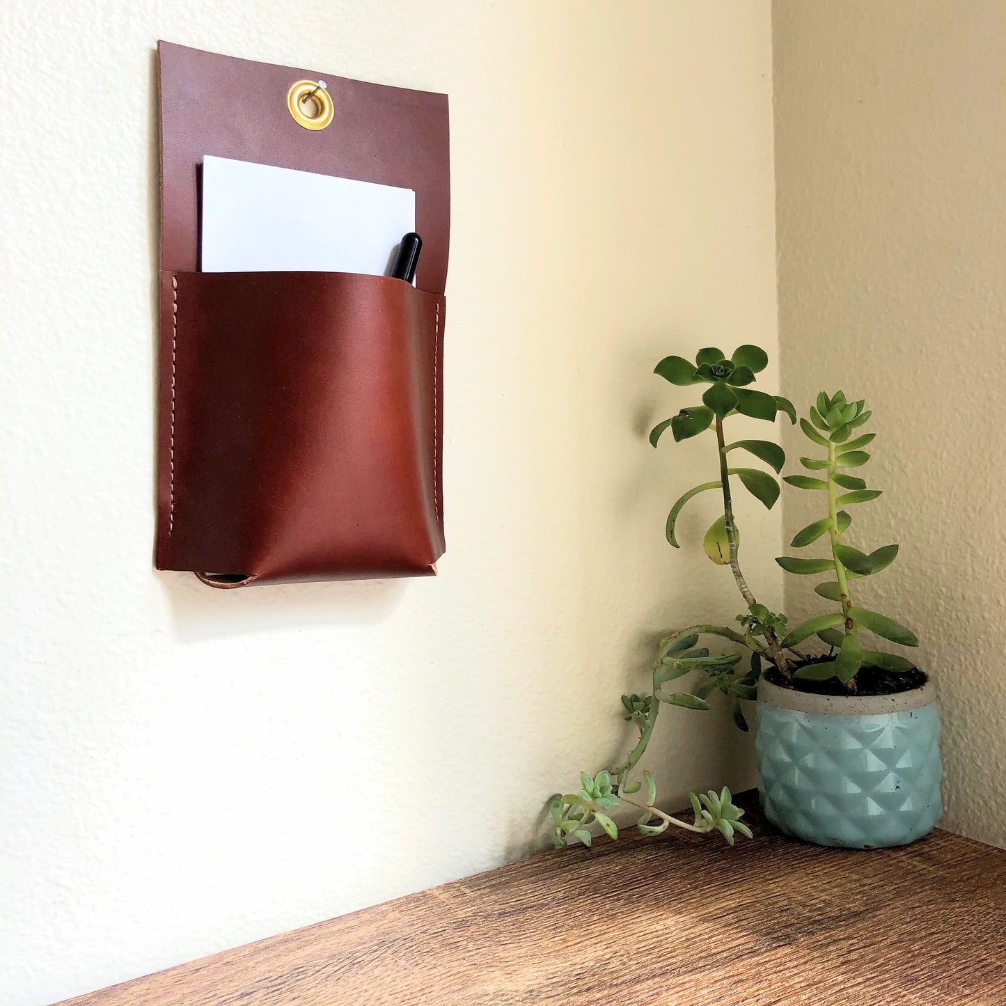 Brown wall caddy holds a pen and paper near a cute potted plant on a table.