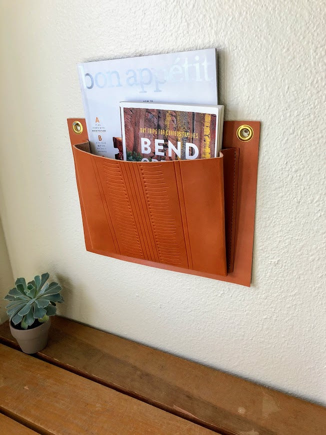 Leather wall pocket with hand tooled geometric design hangs on wall above table and plant.