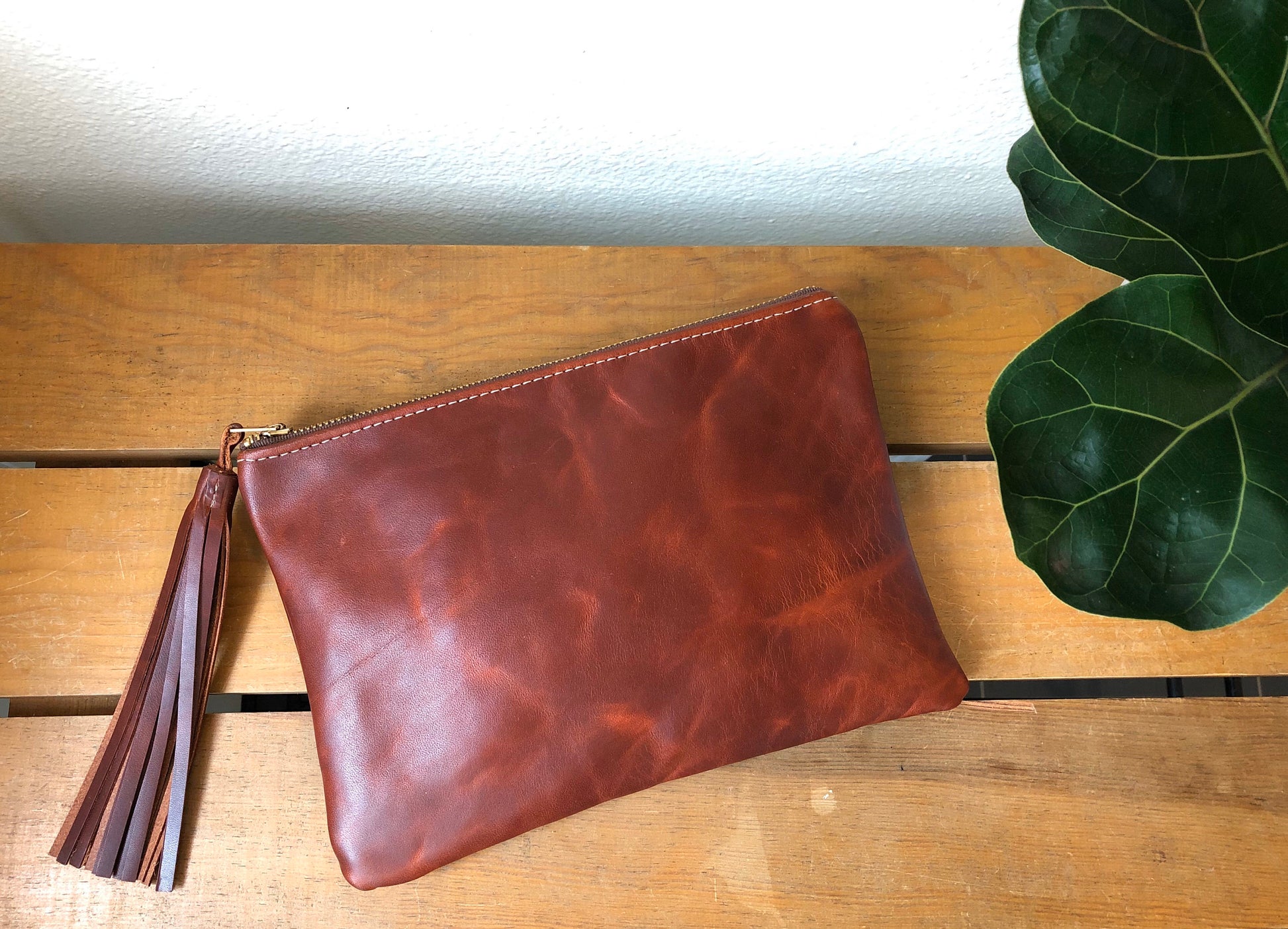 Rich, red brown, tasseled leather clutch lies on wooden table near deep green plant.