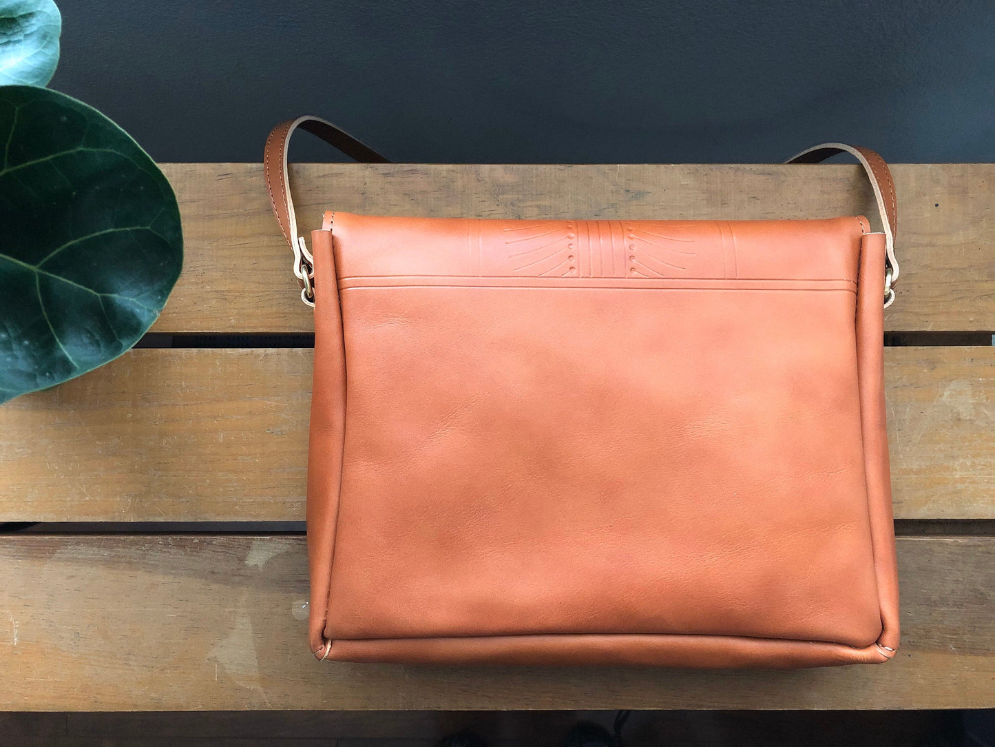 Back of tan leather crossbody bag with small tooling detail. Resting on table near plant