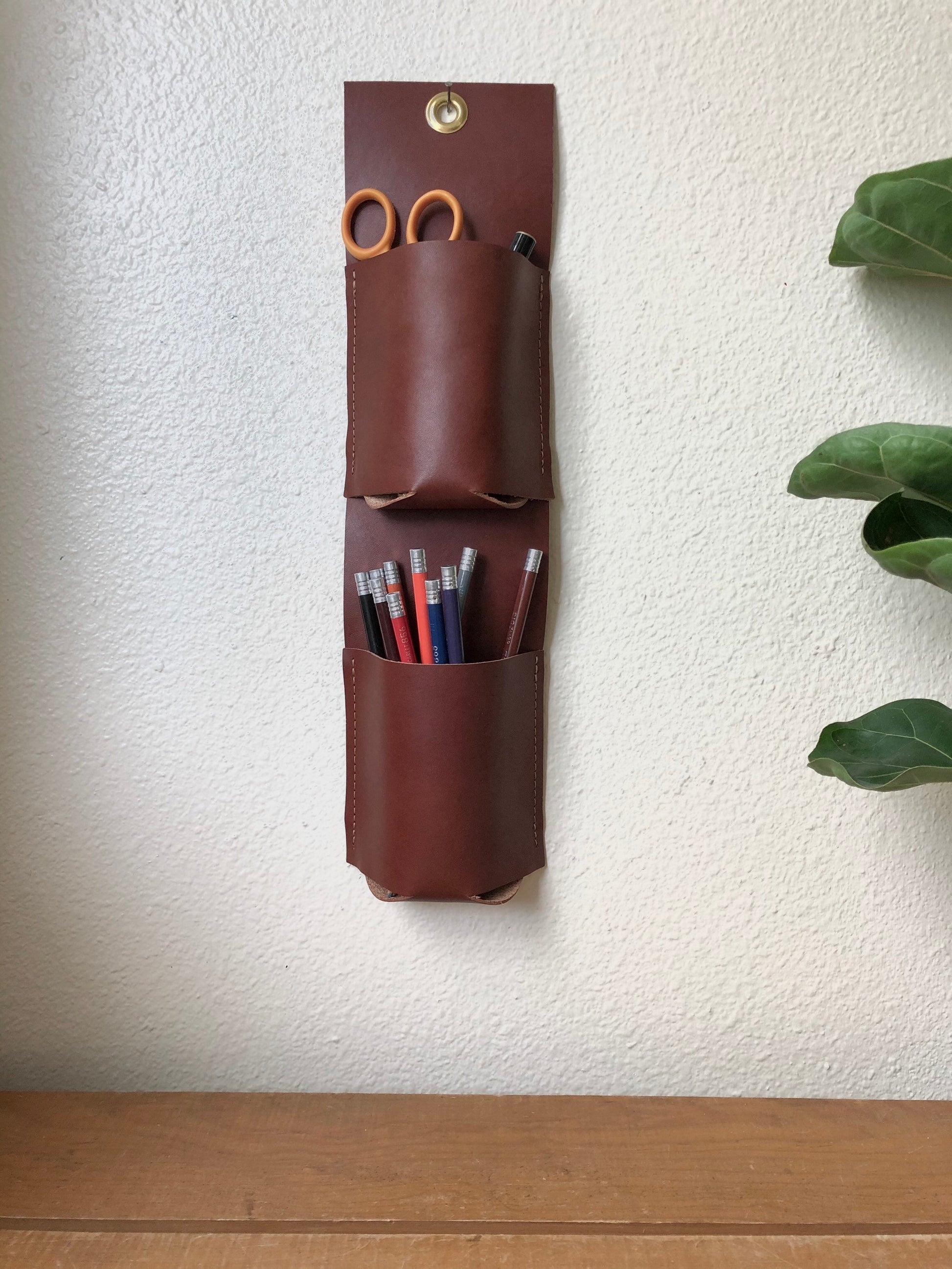 Brown leather hanging organizer with two vertical pockets holds colored pencils, scissors, and pen.