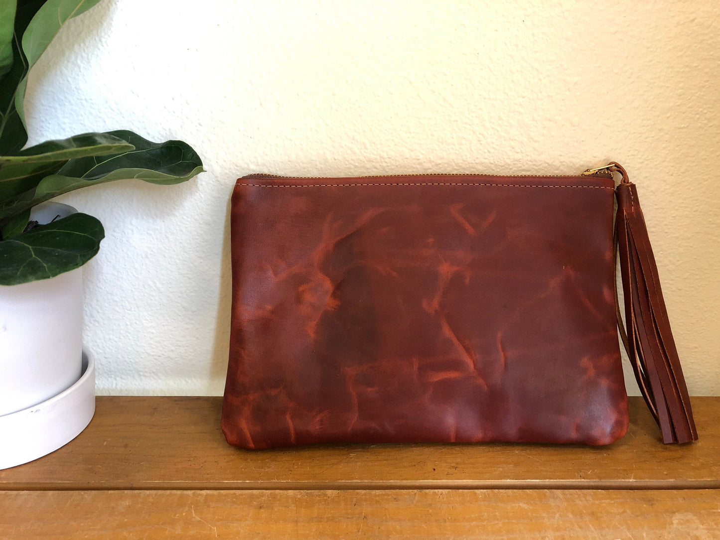 Distressed brown leather clutch with tassel leaning against a white wall on wood table with a plant