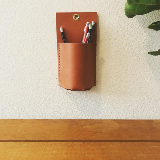 Tan leather wall organizer hangs holding colored pencils above a wood table